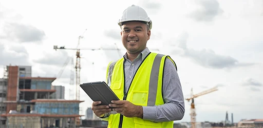 Bauleiter in Sicherheitsweste und Helm mit Tablet auf einer Baustelle im Hintergrund.