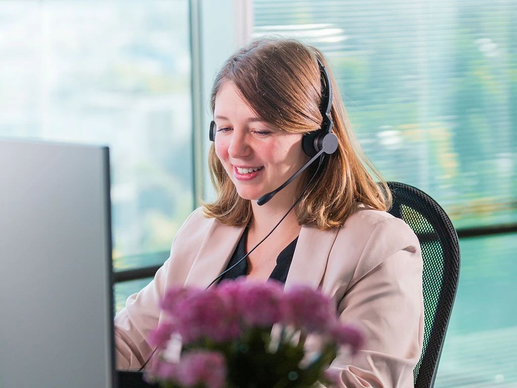 Freundliche Mitarbeiterin im Kundenservice mit Headset am Computer, die lächelnd auf einen Bildschirm schaut.